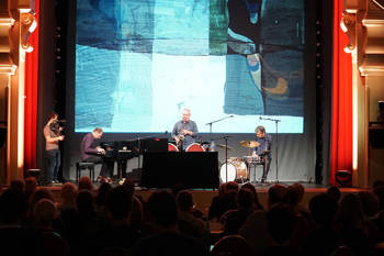 A band consisting of three men playing the piano, the drums, and the saxophone perform on stage